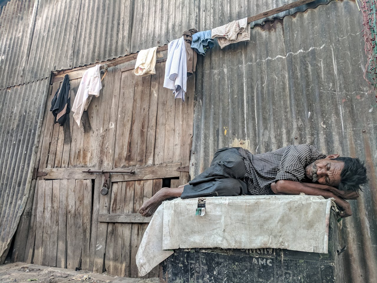 about-us An elderly man peacefully sleeps against a rundown wooden wall with hanging clothes.
