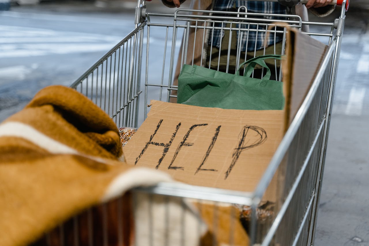 services-01 A shopping cart with a cardboard 'Help' sign and belongings, symbolizing urban homelessness.