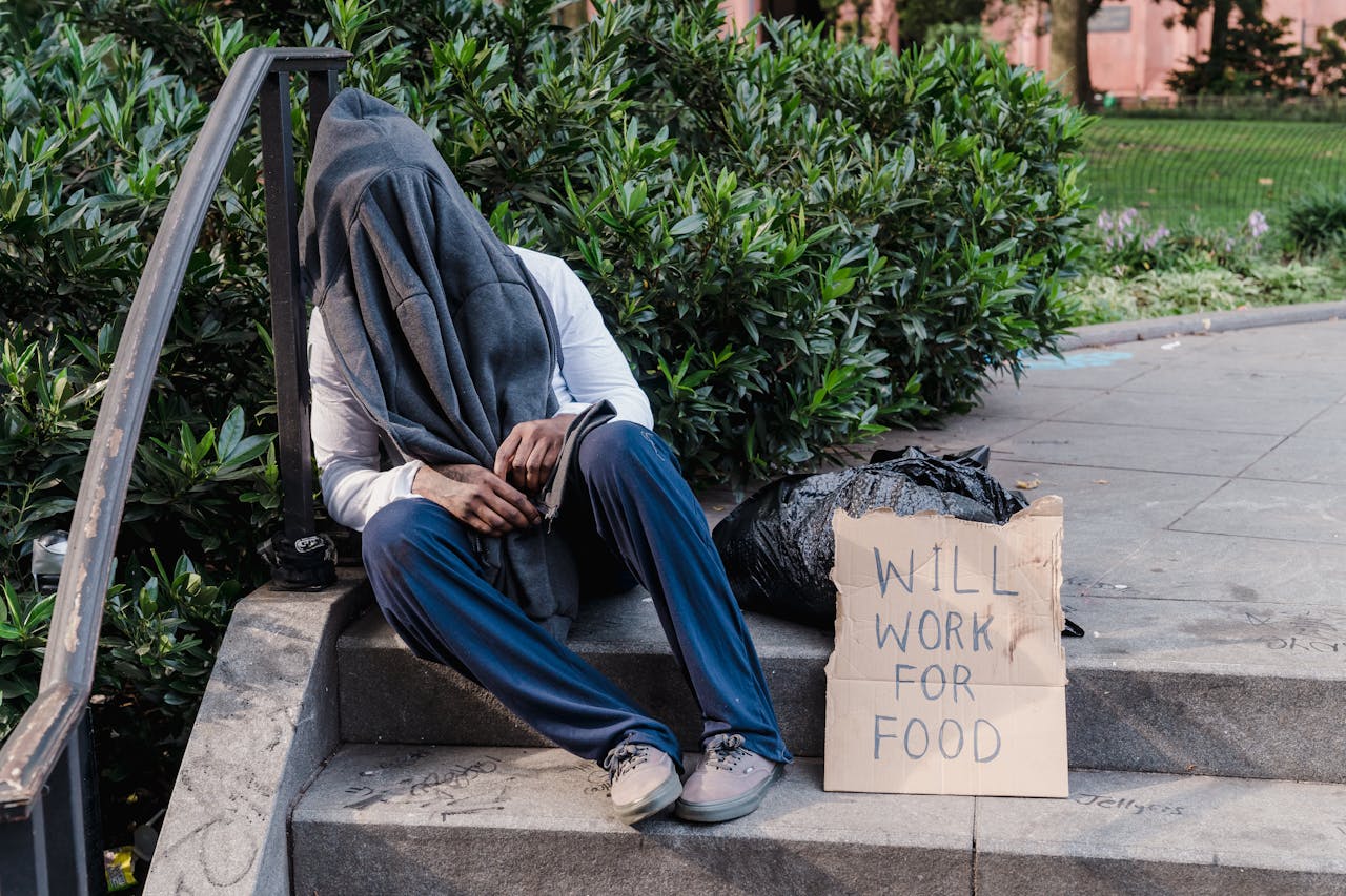 gallery-6 A homeless person sitting on outdoor stairs, covered by a cloth, with a sign reading 'Will Work for Food'.